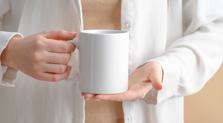 Female hands are holding a mockup of a white empty mug or cup of coffee tea, for your design and advertising, business concept on a beige background, Scandinavian style. Woman in white shirt