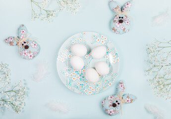 White eggs on plate with spring floral decor, DIY textile Easter bunnies and white flowers on blue.