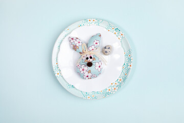 Fototapeta na wymiar Easter festive minimal layout with one quail egg and one DIY textile Easter bunny on plates on light blue backdrop.