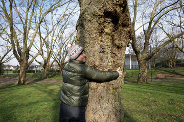 Man wearing green jacket, bobble hat and white pom pom hugging big tree with smiling face in the park in the sunny day