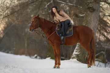 Obraz premium Young woman with long hair in sweater riding a brown horse on snow at sunset. Background of winter trees and sunset sky