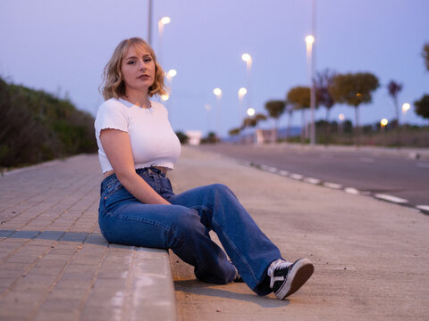 Chica rubia sentada en un bordillo de la carretera por la noche 