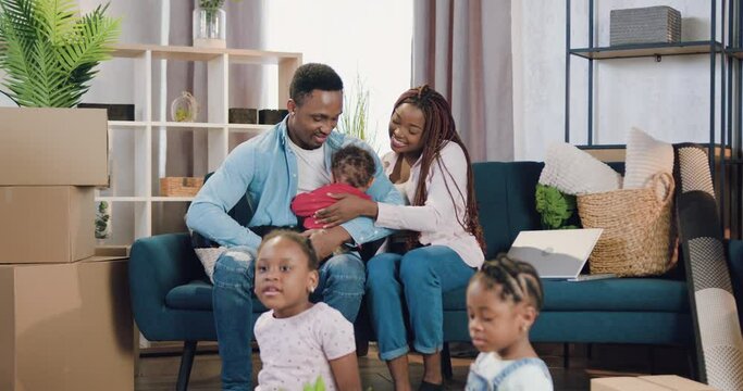 Lovely happy smiling young african american family with small kids moved into newly acquired house and sitting with the smallest baby on the sofa while another two children playing on the floor