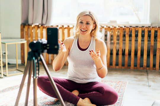 Athletic female fitness and yoga blogger in sportswear recording video of training on mobile phone during workout at home. A middle-aged woman is teaching a video communication lesson and talking.
