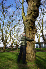 Portrait full body of Caucasian man wearing green jacket, bobble hat and white pom pom hugging big tree with smiling face in the park in the sunny day