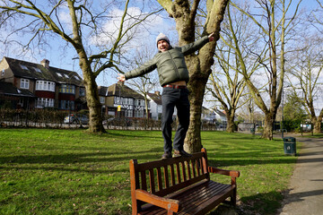 Portrait full body of Caucasian man  wearing green jacket, bobble hat with white pom pom walking on back of wooden bench in the park 