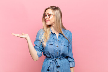 young blond pretty woman feeling happy and smiling casually, looking to an object or concept held...