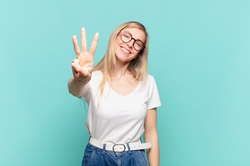 young blond pretty woman smiling and looking friendly, showing number three or third with hand...