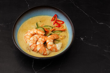 Asian soup with shrimps and vegetables: zucchini, red pepper and carrot. Dish isolated in a blue bowl, close-up on a black marble background. Asian cuisine.