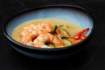 Asian soup with shrimps and vegetables: zucchini, red pepper and carrot. Dish isolated in a blue bowl, close-up on a black marble background. Asian cuisine.
