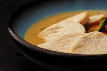 Asian soup with chicken and vegetables: zucchini, red pepper and carrot. Dish isolated in a blue bowl, close-up on a black marble background. Asian cuisine.
