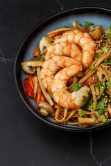 Udon noodles with shrimps and vegetables: zucchini, red pepper, mushrooms, carrot, spring onion and sesame seeds. Dish isolated in a blue bowl, close-up on a black marble background. Asian cuisine.