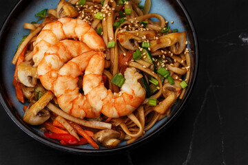 Udon noodles with shrimps and vegetables: zucchini, red pepper, mushrooms, carrot, spring onion and sesame seeds. Dish isolated in a blue bowl, close-up on a black marble background. Asian cuisine.