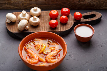 Tom yam soup with shrimps in a tureen on a concrete background next to a bowl with coconut milk mushrooms and tomatoes.