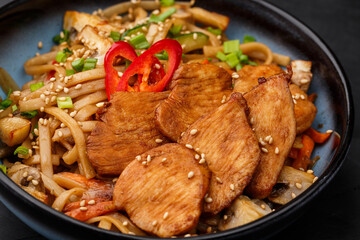 Udon noodles with teriyaki chicken and vegetables: zucchini, red pepper, mushrooms, carrot, onion and sesame seeds. Dish isolated in a blue bowl, close-up on a black marble background. Asian cuisine.