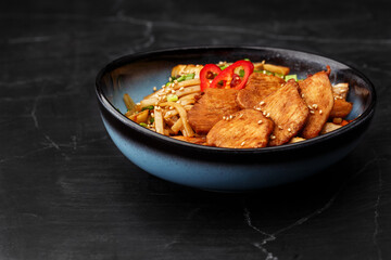 Udon noodles with teriyaki chicken and vegetables: zucchini, red pepper, mushrooms, carrot, onion and sesame seeds. Dish isolated in a blue bowl, close-up on a black marble background. Asian cuisine.