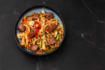 Udon noodles with teriyaki beef and vegetables: zucchini, red pepper, mushrooms, carrot, onion and sesame seeds. Dish isolated in a blue bowl, close-up on a black marble background. Asian cuisine.