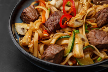 Udon noodles with teriyaki beef and vegetables: zucchini, red pepper, mushrooms, carrot, onion and sesame seeds. Dish isolated in a blue bowl, close-up on a black marble background. Asian cuisine.