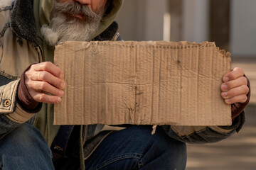 Senior man homeless beggar with a gray beard in a shabby clothes with a .carboard sign sitting...