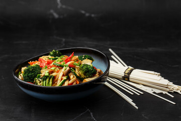 Udon noodles with vegetables: zucchini, broccoli, red bel pepper, mushrooms, carrot, spring onion and sesame seeds. Dish isolated in a blue bowl, close-up on a black marble background. Asian cuisine.
