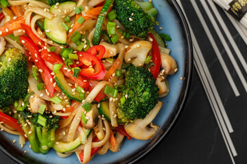 Udon noodles with vegetables: zucchini, broccoli, red bel pepper, mushrooms, carrot, spring onion and sesame seeds. Dish isolated in a blue bowl, close-up on a black marble background. Asian cuisine.