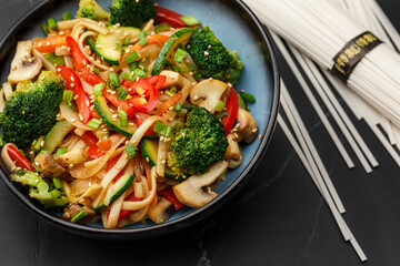 Udon noodles with vegetables: zucchini, broccoli, red bel pepper, mushrooms, carrot, spring onion and sesame seeds. Dish isolated in a blue bowl, close-up on a black marble background. Asian cuisine.
