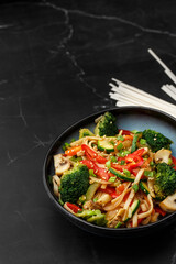 Udon noodles with vegetables: zucchini, broccoli, red bel pepper, mushrooms, carrot, spring onion and sesame seeds. Dish isolated in a blue bowl, close-up on a black marble background. Asian cuisine.
