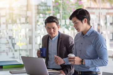 Two Asian businessman are meeting for a collaboration concept using a laptop  holding coffee  at the office.