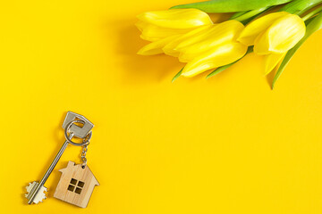 Key ring in the shape of wooden house with key on yellow background and spring tulips. Building, design, project, moving to new home, mortgage, rent and purchase real estate, summer offer. Copy space