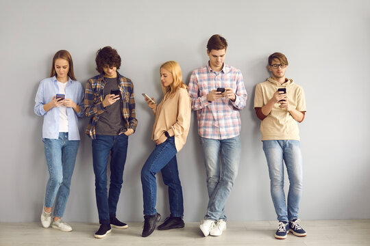 Group Of Young People Standing Leaning On Wall, Using Cell Phones, Chatting Online, Scrolling Through Social Media Feed And Ignoring Each Other. Mobile Device Addiction, Excessive Gadget Use Concept