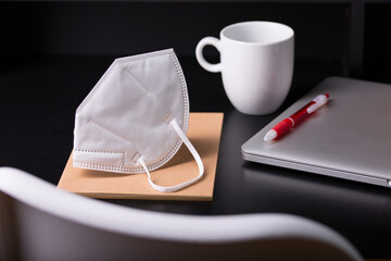 Close up view of a black work desk interior with a laptop computer, a cup of coffee,  paper, pen, plant, face mask for coronavirus black background