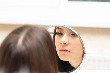 Fototapeta premium Young woman putting on makeup while looking in the mirror paints eyelashes mascara for the eyes