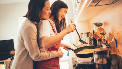 Fototapeta na wymiar Happy Lesbian Couple Cooking Together in the Kitchen. Girlfriend Embracing Her Partner from the Back while they Prepare Preparing Delicious Meal. Young Partners in Love, Talking, Having Fun. 