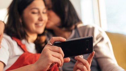 Happy Lesbian Couple Resting on the Sofa Use Smartphone. Sunny Morning Two Girlfriends in Love Have Fun Kissing, Watching Videos, Doing e-shopping. Young Partners Share Tender Moments