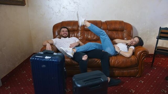Relaxed man and woman waiting for the departure. Young couple in casual clothes laying on a sofa. Packed suitcases on floor. Finish the vacation.