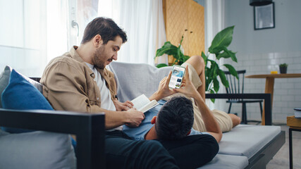 Happy Gay Couple Spending time Together at Home. Partner Using Smartphone and Lying with head on...