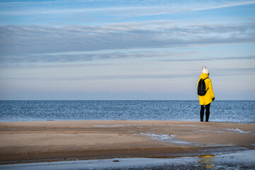 Obraz premium A woman in a yellow winter jacket and with a black backpack walking by the shores of the Baltic Sea near Carnikava, Latvia