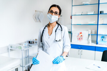 Female doctor in a protective mask and with a stethoscope, holding documents in the middle of a modern hospital. Medicine, health and self-care