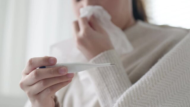 Asian woman checking body temperature by digital thermometer on sofa at home. Self isolation at coronavirus covid-19 quarantine,having fever. Infected patient at home medical self treatment.