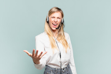 young blond telemarketer woman looking angry, annoyed and frustrated screaming wtf or what’s...