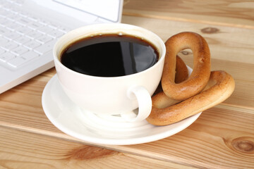A white cup of coffee or chicory and drying on a saucer on a wooden table next to a white laptop.