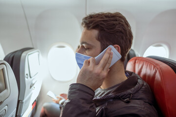 one male person with mask talks to phone on plane seat before taking off and sun shines from aircraft window