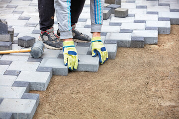 A worker wearing yellow protective gloves puts paving slabs on a prepared flat sandy pad on the...