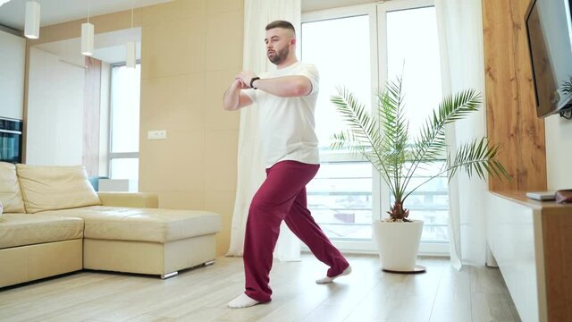 young bearded handsome man doing physical exercises on squats in pajamas at home in the living room. Male workout for weight loss in the house. training, bodycare and wellness concept