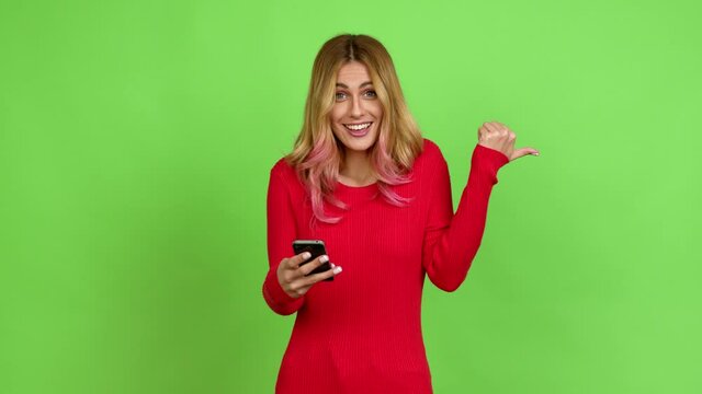 Young blonde woman using mobile phone and pointing side over isolated background on green screen chroma key