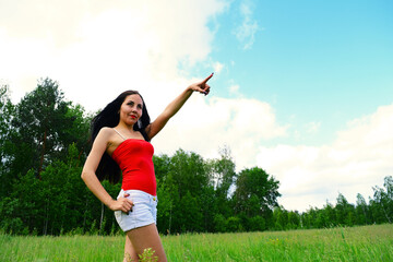 Fototapeta na wymiar Young woman in a red t-shirt and denim shorts shows a finger to the sky while standing on nature near the forest