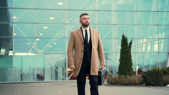 young stylish bearded business man walks down the city street in a suit, coat and briefcase in hands. on urban background of a modern building. Portrait a successful and confident businessman walking