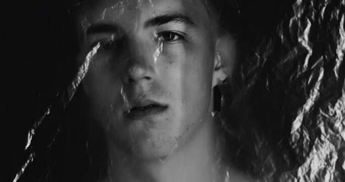 Man discrimination. Social pressure. Fear isolation. Defocused black and white portrait. Depressed scared guy face hiding behind wrinkled ripped polyethylene film on dark background out of focus.