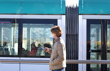 Young  man wearing  surgical face mask waiting for the train,using smartphone, typing a message on the phone.
