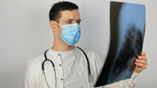 A young handsome doctor in a medical mask holds an x-ray picture of the lungs in his hands and conducts an analysis. A young surgeon wearing a protective medical mask examines an X-ray of a patient's